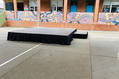 Outdoor stage platform in a school courtyard with black skirting, side access step and a colourful mural backdrop