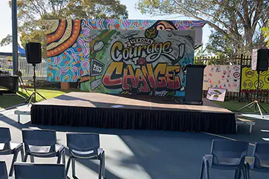 School event stage with black skirting, a lectern, speakers and audience seating in front of a colourful mural wall