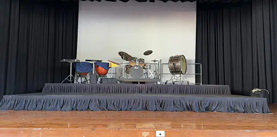School performance stage with black skirted platforms, a raised drum riser, percussion instruments and theatre curtains on a wooden hall floor