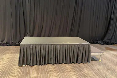 Small stage podium with black skirting, access step and black curtain backdrop inside an indoor event room with patterned carpet