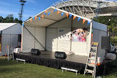 Stage cover hire setup with a white marquee roof, black skirted performance platform, speakers, access steps and event signage beside a stadium