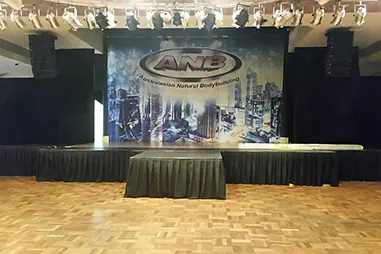 Stage hire setup for a bodybuilding event with overhead lighting, black platform panels and a large branded backdrop behind the competitors