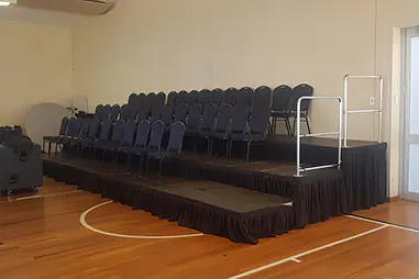 Tiered event seating with black modular risers, rows of padded chairs, safety handrails and black stage skirting inside an indoor hall