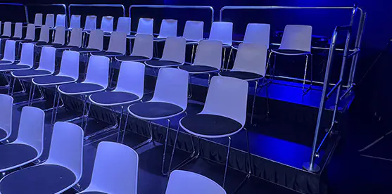 Tiered event seating with white chairs on black risers, safety rails and blue venue lighting in a dark indoor auditorium