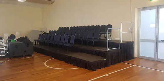 Tiered staging hire with black modular risers, rows of padded black chairs, safety handrails and black stage skirting inside an indoor hall
