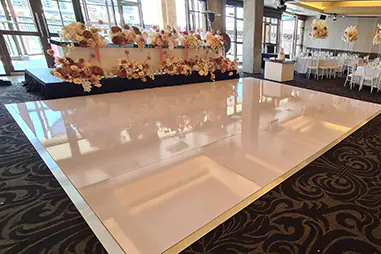 Wedding reception stage and white dance floor beside formal dining tables, white chairs and tall floral centrepieces in a large indoor venue