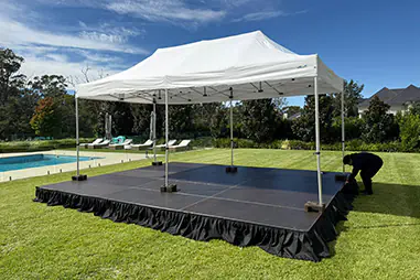 White canopy stage platform with black skirting set up on a lawn beside a swimming pool, garden beds and outdoor entertaining area under a clear blue sky