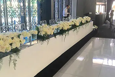 White wedding stage hire setup with a long bridal head table, glossy white stage panels, white floral arrangements, clear chairs and black trim in an elegant indoor venue